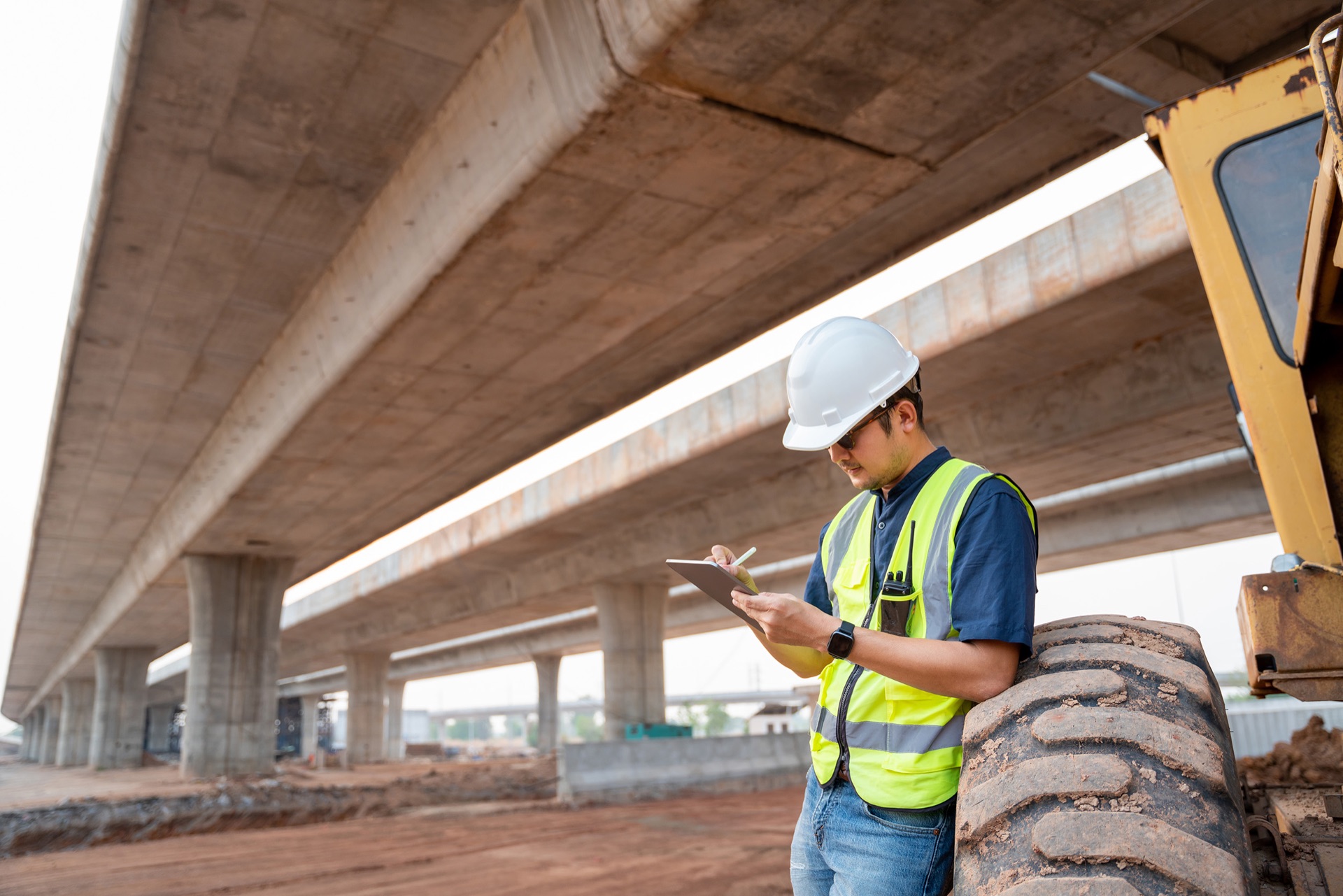 An inspector in a hard hat and hi-vis recording checks on a clipboard beside heavy equipment